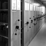 Black-and-white image of a compact archive storage system with numbered shelving units and hand-crank mechanisms, symbolizing classification, knowledge organization, and institutional control.