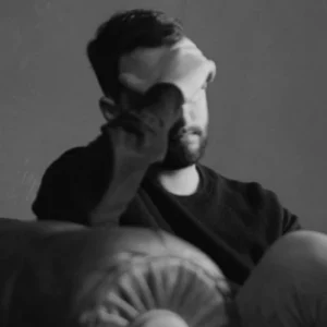 Young man sitting alone on a couch holding his head, expressing stress, frustration, and emotional strain related to General Strain Theory in criminology.